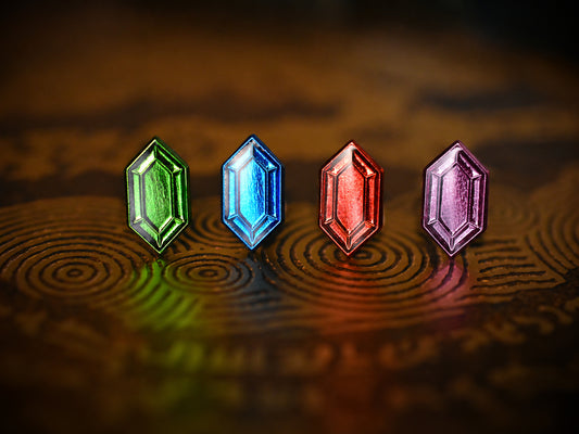 Four miniature jewel-shaped enamel pins in metallic green, blue, red and violet purple arranged on an engraved fantasy-inspired surface.